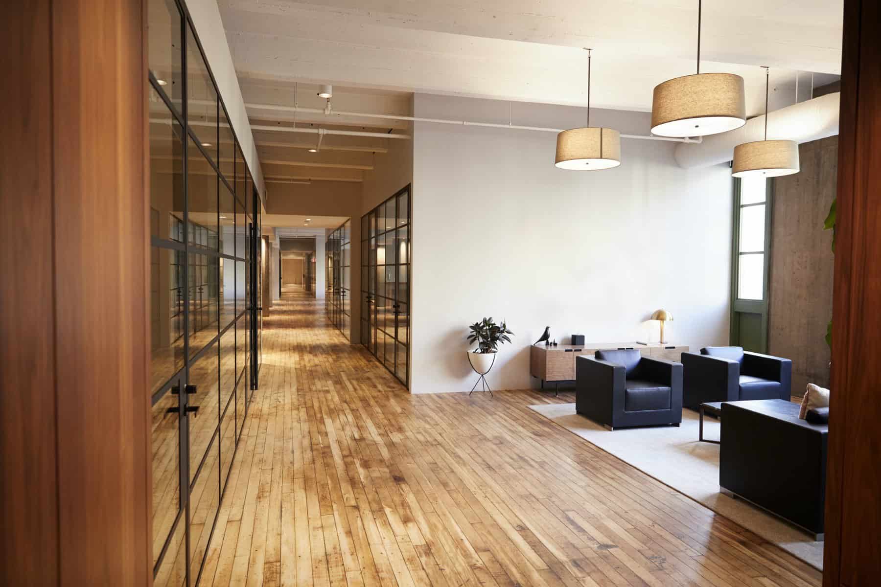 Bright commercial interior hallway with white painted walls, wood-framed glass partitions, warm wood flooring, and pendant