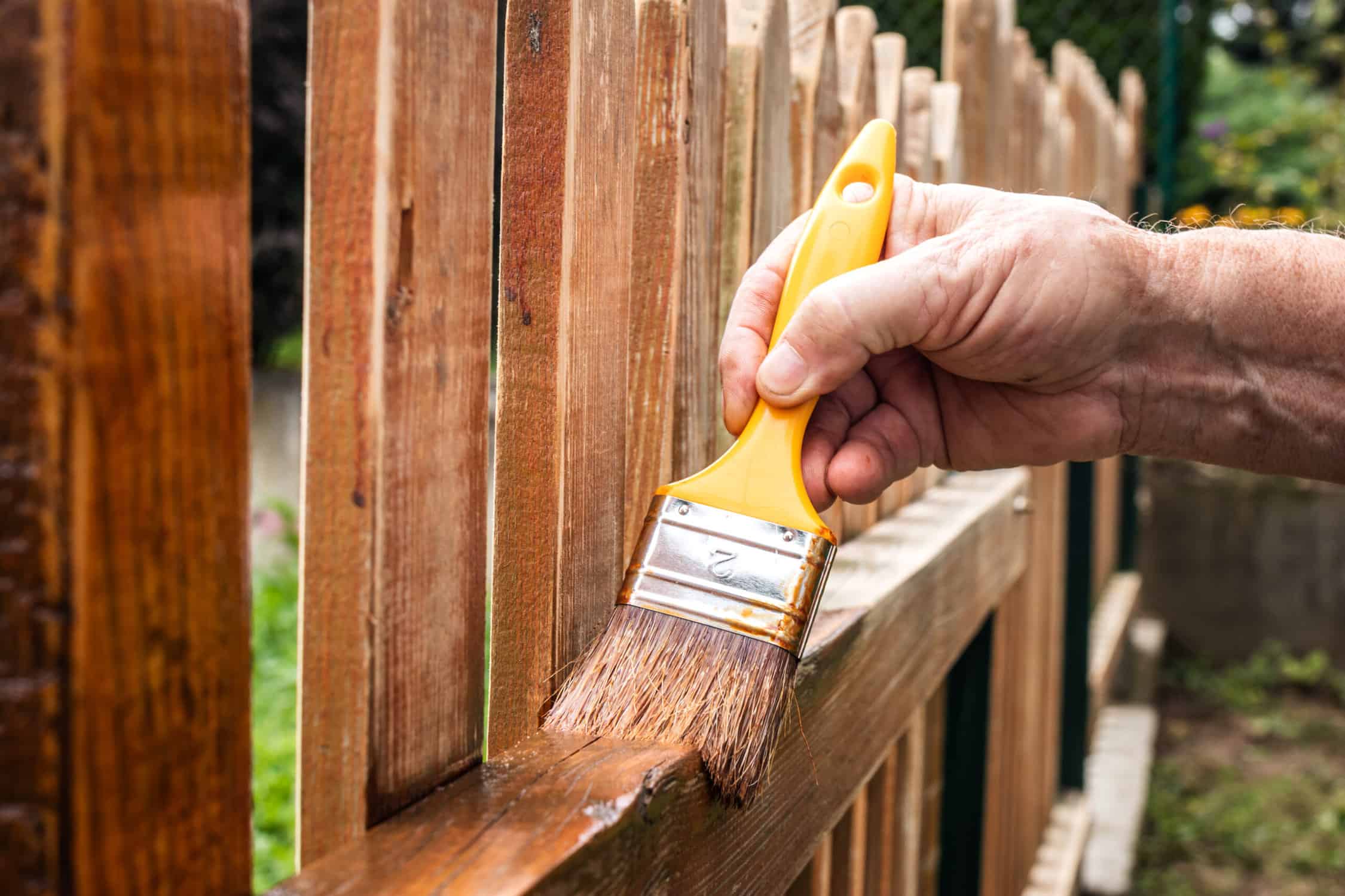 Wood Fence Stain Application Worker applying golden-brown wood stain to weathered fence boards with yellow-handled paintbrush.
