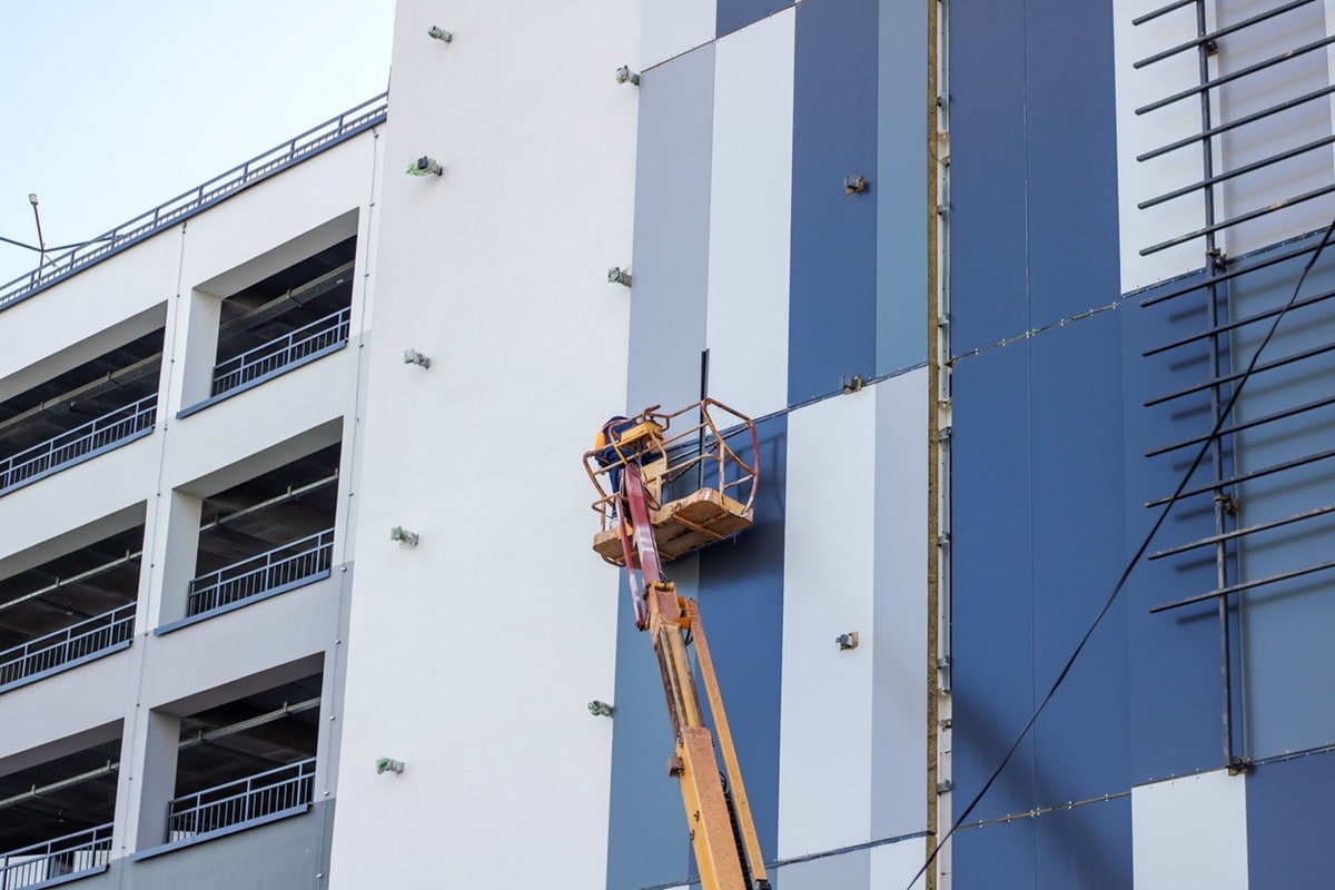 A man inside a lift is painting the side of a building A man is seen inside a lift painting the side of a building. The building is a tower block or condominium in an urban setting. A crane and electricity lines can be spotted nearby