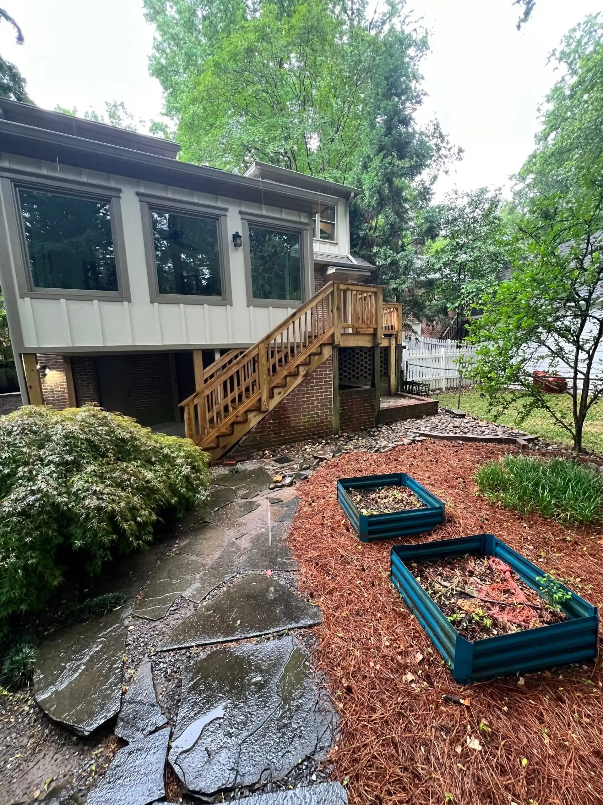 Exterior Residential Home Before White and gray exterior residential home with wooden deck stairs, brick foundation, and reddish-brown mulch landscaping in